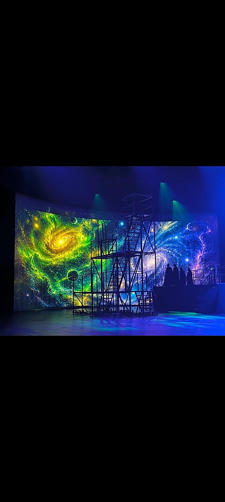 Bühnenbild mit Galaxie-Projektion und Darstellern auf einem Gerüst