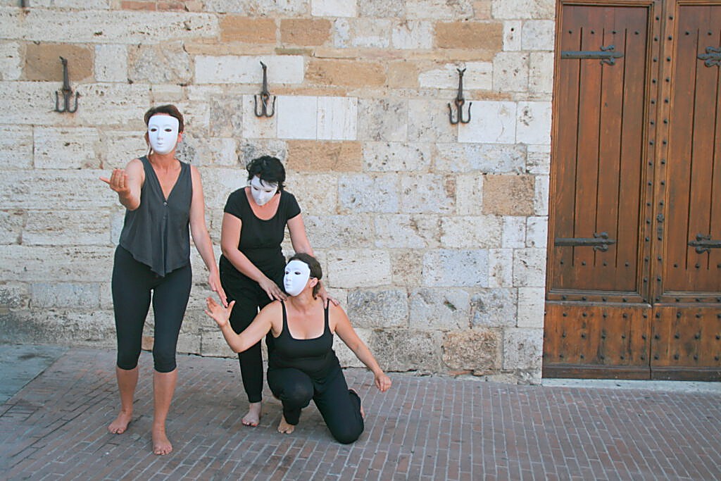 Theatergruppe mit weißen Masken vor einem historischen Holztor